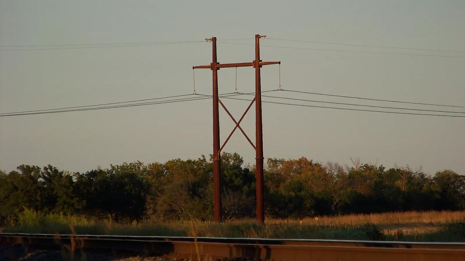 A steel H-frame transmission structure at dusk beside a railway, with trees and grassland behind