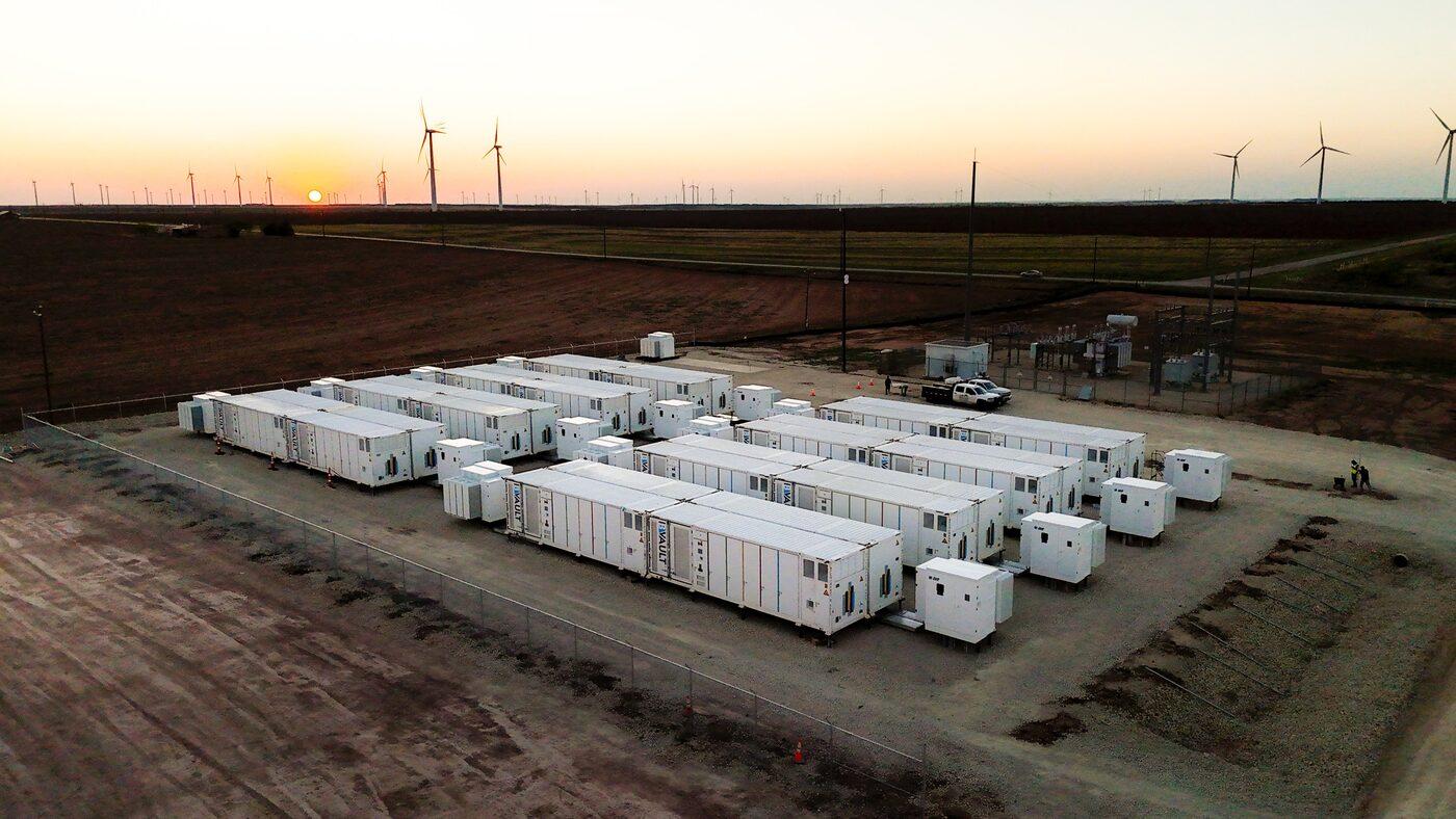 Drone photograph at sunset of the Cross Trails battery storage site in Scurry County, Texas: rows of white battery containers on a gravel pad with wind turbines on the horizon.