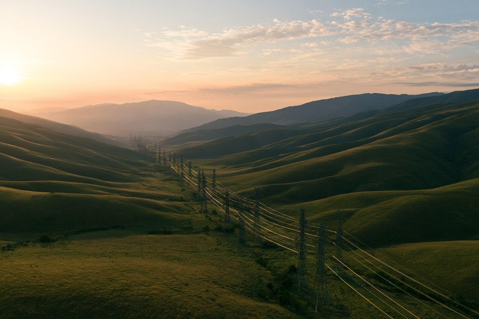 A high-voltage transmission corridor cutting across rolling green hills at dawn
